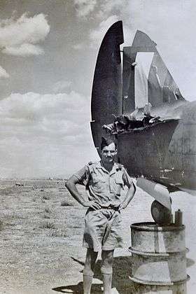 Man in jhaki shirt and shorts, wearing forage cap, beside aircraft missing part of tailplane