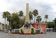 A tall sweeping stone triangle projects skyward behind the statues of a man and two children in bronze on a smaller podium. Around the base are placed several wreaths with logos. Palm trees surround the scene.