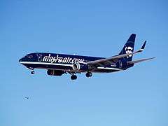 Left side view of an Alaska Airlines 737-800 painted in a  primarily black livery, and says "alaskaair.com" on both sides. Aircraft is airborne.