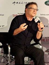 A man wearing a black shirt and trousers, sitting on a chair and talking through a microphone.