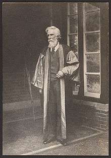 A bearded Albert Venn Dicey stands in a suit and academic robes facing left