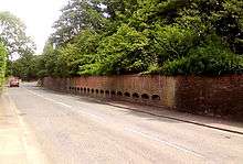 Photograph of a wall with 17 semi-circular grilles built into it. The grilles are spaced horizontally along the wall's length