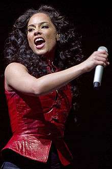 The photo shows an African American female with medium brown hair with her head tilted to her right with her mouth open. She is wearing a red sleeveless shirt with dark wash colored jeans and is holding a microphone with her right hand.