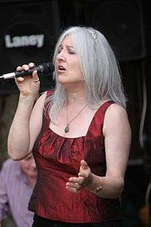 Silver-haired woman wearing a crimson sleeveless top and holding a microphone
