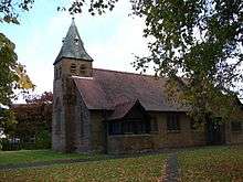 A small church with a tower and broach spire to the left, trees are to the left and overlapping to the front and autumn leaves lie on the grassed area in the foreground