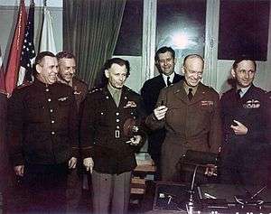 Group shot of six smiling men (and one barely visible woman), each in a different military uniform, standing behind a writing desk. Eisenhower is holding three fountain pens. In the background are flags, including the US flag.