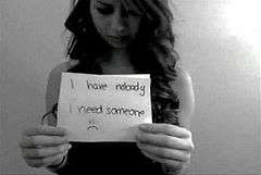 A girl in her mid-teens holding a cut-out piece of paper or card with the words "I have nobody, I need someone" and a sad emoticon written on it.