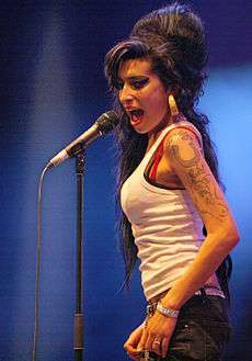 Amy Winehouse performing on 29 June 2007 at the Eurockéennes music festival in eastern France. Winehouse is looking into the crowd while singing sidelong into a handheld microphone. Her black hair is combed back in a relaxed style. Her tattooed shoulder is bare, and she is wearing large, chunky earrings.