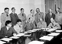A dozen men behind a large table, half of whom are in military uniform and half in civilian clothes, seven standing and five seated