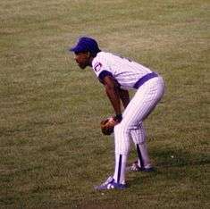 African-American man in a white baseball jersey with blue trim standing bent at the waist on a green field.