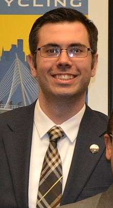 A white man with brown hair and glasses and a suit smiles widely at the camera.