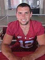 A white man with close-cropped dark hair sits at a table, looking into the camera. The man is wearing a football jersey emblazoned with the number 12.