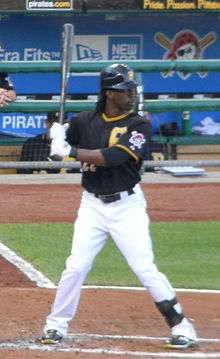 A dark-skinned man in a black baseball jersey and cap, white pants, and white batting gloves stands in a right-handed batting stance.