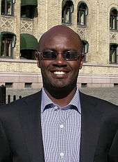 A man in a suit and sunglasses, Andrew Mwenda, smiling