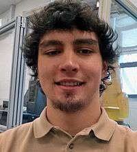 A young Caucasian man wearing a light brown collared shirt with dark brown curly hair and a slight beard on the chin