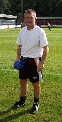A middle-aged man, wearing a white T-shirt and black shorts, standing on a grass field.
