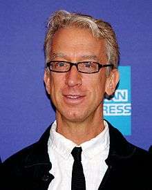 A smiling Andy Dick with glasses, wearing black and white clothes with an American Express logo behind him