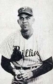 A black-and-white photograph of a man wearing a white pinstriped baseball jersey with "Phillies" across the chest and a dark undershirt; he is holding a baseball in his lap with both hands and wears a baseball cap with a white "P" on the face atop his head