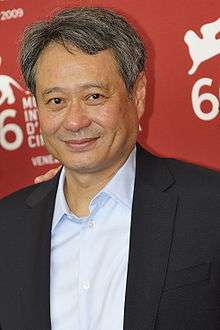 An Asian male wearing a grey jacket over an unbuttoned blue shirt is standing in front of a red wall with white text.