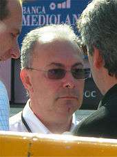 A man in his fifties, visible only from the neck up, wearing glasses with tinted lenses and a white polo shirt. Two other men are partly visible on either side of him.