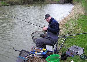 Man seated at the side of the water surrounded by fishing rods and tackle.