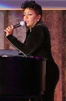 A woman sings while she is recharged on a piano. She wears a long black ensemble and diamond earrings.