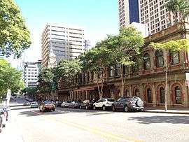 view of Ann Street in sunshine with a few travelling cars, parked cars in front of Central railway station