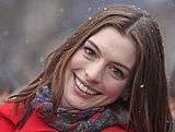 A photograph of Anne Hathaway; her head is tilted sidewards and she is wearing a red overcoat with a purple scarf.