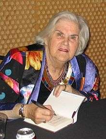 A woman with white hair wearing a light black and multicoloured jacket is seated at a table signing a book