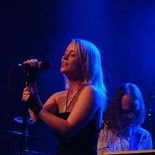 Annie performing behind a microphone stand with a keyboardist in the background.