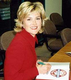A colour photograph of a woman with blonde hair and blue eyes wearing a red dress shirt, writing in a book, and smiling at the viewer