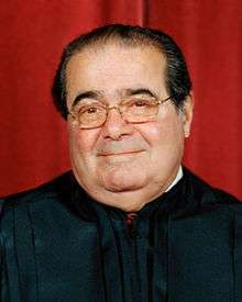 A photographic portrait of the head and upper body of a man with a receding dark hairline, wearing glasses and a black robe