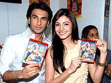 Anushka Sharma and Ranveer Singh holding up DVDs. While Sharma, wearing a pale yellow top is smiling directly at the camera, Singh, dressed in a white shirt, is looking away.