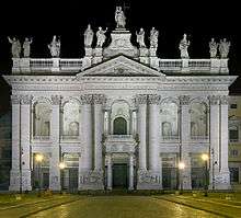 Ornate facade of the basilica at night with columns, main door, and statues of the twelve Apostles on the roofline, with a Latin inscription below them