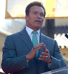 Man wearing a blue suit and standing at a podium. He is facing away from the camera, smiling, and is clapping.