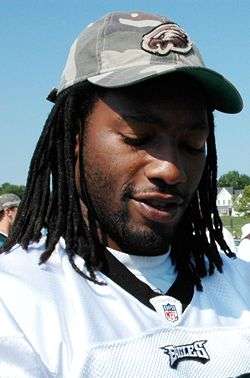 A African-American man with neck-length dreadlocks and wearing a jersey and baseball cap looks down.
