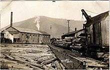 Black and white image shows a large building with a smokestack at left and many large logs in the foreground. At right is a logging train with a loader crane on a car. A bare mountain is in the background.