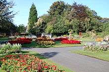 Pathway leading towards a circular flowerbed where the path splits. Flowerbeds also flank the path on each side. The flowerbeds are decorated by various red and yellow flowers. Trees are in the background.