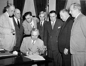A man in a suit is seated at a desk, signing a document. Seven men in suits gather around him.