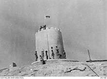 Soldiers stand at the base of a sandstone structure in the desert whilst a flag is unfurled on top of the building