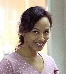 An Indonesian woman, looking forward and smiling. She is wearing a purple v-neck shirt