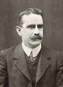 a formal black and white portrait of a male with a moustache wearing a suit