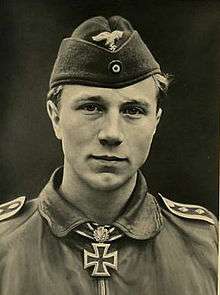 The head and shoulders of a young man, shown from the front. He wears a field cap and a military leather pilot jacket, with an Iron Cross displayed at the front of his collar. His facial expression is a determined and confident smile; his eyes are looking into the camera.