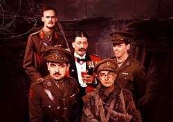 The main members of the cast of Blackadder Goes Forth are seated in the production's World War I trench set. Seated in the centre is Stephen Fry as General Melchett, incongrously wearing red officer's mess dress and holding a sherry. Tim McInnerny as Captain Darling and Hugh Laurie as Lieutenant George stand beside him in green officers' uniforms. Most prominent in the picture is Rowan Atkinson as Captain Blackadder, wearing his captain's uniform. To his right is Tony Robinson as Baldrick, wearing a moth-eaten private's uniform, his cap angled off-centre. George is the only one smiling.