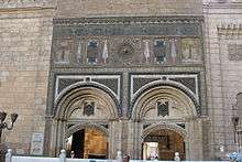 Two large arches recessed into a wall are visible, with entrance-ways at their bottom. Above the arches the wall is ornately decorated with carvings and geometric designs. Parts of the decorations are colored red, blue and gold. The arches and decorations are surmounted and flanked by large, closely set rectangular blocks.