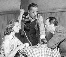 Lauren Bacall in black and white sitting at a table with Humphrey Bogart and Henry Fonda while performing a scene on an episode of The Petrified Forest in 1956