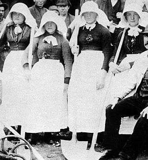 Four women wearing dark heavy clothing, bright white aprons, and long white bonnets entirely covering the sides of their heads and protruding forwards over their faces