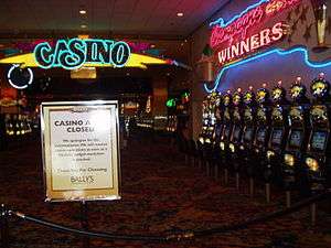 A roped off entrance to room with a neon sign "CASINO", over a sign saying "CASINO A... CLOSED". The room contains many slot machines. Nobody is in the room.