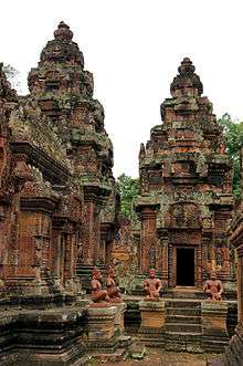 Ancient Cambodian open-air temple, featuring four statues of seated figures in the middle distance.