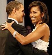 Barack and Michelle Obama dance arm-in-arm and smile. She wears a white dress, large ring, long earrings and a bracelet. He wears a black tuxedo.